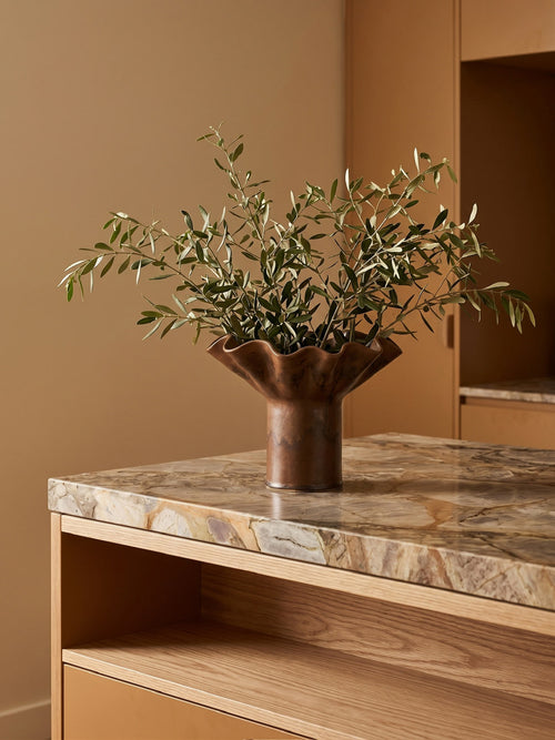 A sculptural chocolate-toned vase with a flared, scalloped rim holding green olive branches, placed on a marble-topped wooden console in a warm, minimalist interior.