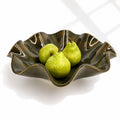 An oily Rockpool coloured wavy ceramic bowl with a glossy glaze, containing three green pears, set against a clean white background with soft window shadow patterns.