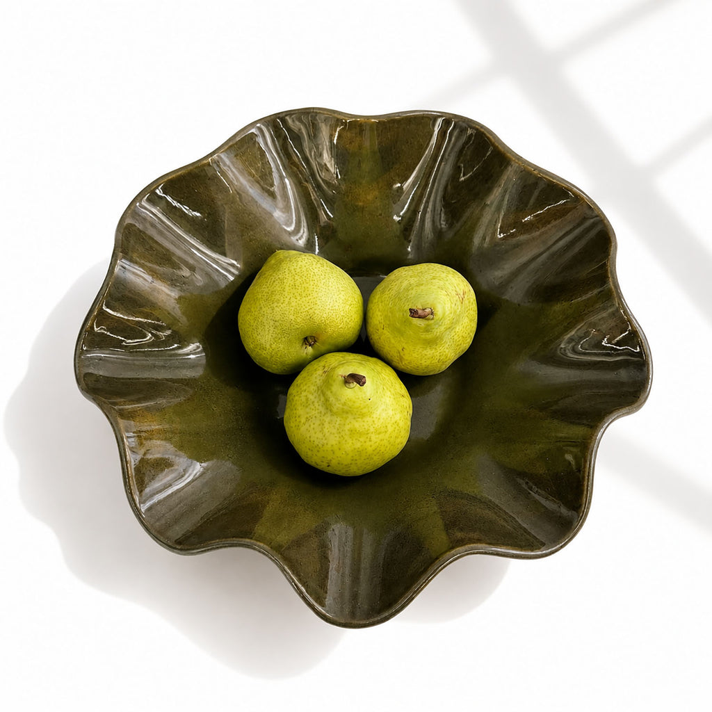 A wavy ceramic bowl with a glossy dark olive-green glaze, containing three green pears, photographed from above on a white background with soft window-light shadows.
