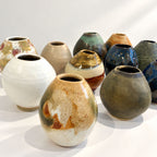 A group of handmade ceramic vases in various earthy glazes, including white, brown, blue, green, and terracotta, arranged on a white surface against a white background.