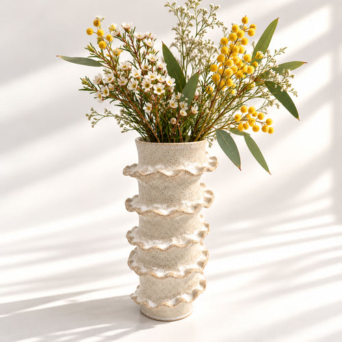 A textured cream-coloured ceramic vase with a stacked, wavy tiered design, filled with a floral arrangement of yellow berries, white blossoms, and green leaves